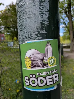 Street sticker A vibrant rectangular sticker with a bright green background, featuring a collection of elements associated with Hammarby IF and Södermalm, Stockholm. On the left, a stylized golden head wearing a crown is depicted. To its right, a large, domed building (resembling Avicii Arena) is shown in grayscale, with the 'Medborgarplatsen' subway station entrance visible below it. Further right, a tall, cylindrical building with a distinctive top (likely Katarinahissen) stands in grayscale. A cartoonish figure, dressed in green and holding the yellow and green circular emblem of Hammarby IF, is positioned in front of the buildings. The bottom of the sticker features prominent white text with a dark outline: 'BAJEN FRÅN' in smaller letters above 'SÖDER' in large, bold letters. A thin white border and a light blue strip are visible at the very bottom edge of the sticker.