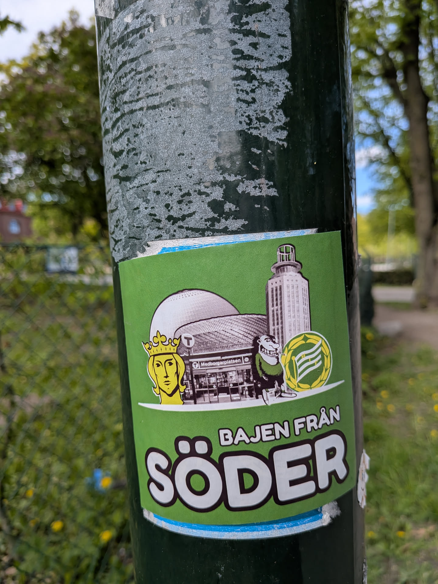 A vibrant rectangular sticker with a bright green background, featuring a collection of elements associated with Hammarby IF and Södermalm, Stockholm. On the left, a stylized golden head wearing a crown is depicted. To its right, a large, domed building (resembling Avicii Arena) is shown in grayscale, with the 'Medborgarplatsen' subway station entrance visible below it. Further right, a tall, cylindrical building with a distinctive top (likely Katarinahissen) stands in grayscale. A cartoonish figure, dressed in green and holding the yellow and green circular emblem of Hammarby IF, is positioned in front of the buildings. The bottom of the sticker features prominent white text with a dark outline: 'BAJEN FRÅN' in smaller letters above 'SÖDER' in large, bold letters. A thin white border and a light blue strip are visible at the very bottom edge of the sticker.