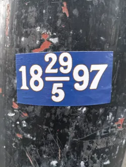A blue sticker with white numbers, reading "18 29/5 97", is affixed to a weathered, dark-colored pole. The numbers are large and bold, suggesting a possible code or reference. The sticker is rectangular and shows some minor wear from its placement outdoors.
