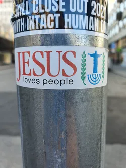 A sticker expressing a message of love and faith. It features the word 'JESUS' in large, orange letters, followed by the phrase 'loves people' in a smaller font.  A blue image of Christ the Redeemer statue is present alongside wheat stalks and a menorah, suggesting a unifying message of faith. The sticker is affixed to a metallic pole.