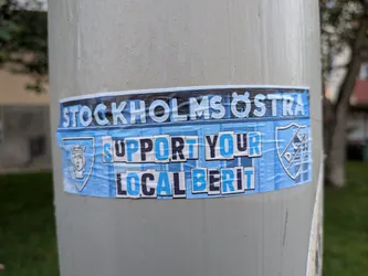 Street sticker The sticker features a predominantly blue and white color scheme. The top band of text reads 'STOCKHOLMS ÖSTRA' in bold white letters with a thin black outline, against a dark blue background with subtle horizontal lines. Below this, the main message is presented in two lines: 'SUPPORT YOUR' and 'LOCAL BERIT'. These words are also in bold, alternating between blue and white letters with black outlines, set against a textured light blue background that mimics crumpled paper. On the left side of the sticker, there's a shield emblem with a stylized portrait of a crowned female figure in white. On the right side, a circular emblem with the letters 'D.I.F.' is visible, also in white within a blue outline. The overall appearance suggests a fan-made or promotional sticker, likely for a sports team or local initiative associated with Stockholm Östra and the name 'Berit'.