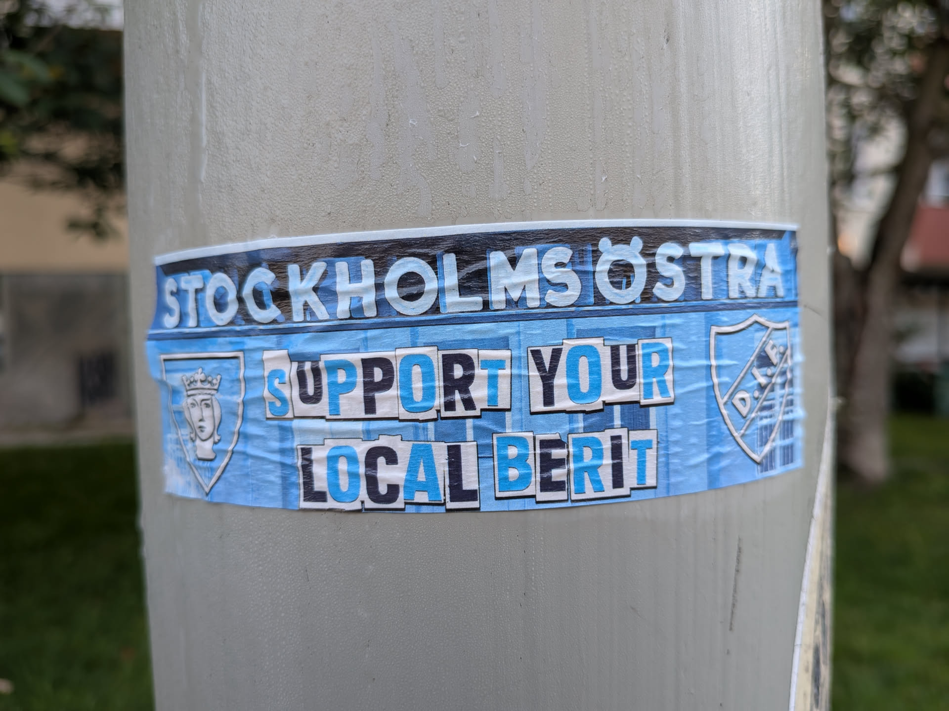 The sticker features a predominantly blue and white color scheme. The top band of text reads 'STOCKHOLMS ÖSTRA' in bold white letters with a thin black outline, against a dark blue background with subtle horizontal lines. Below this, the main message is presented in two lines: 'SUPPORT YOUR' and 'LOCAL BERIT'. These words are also in bold, alternating between blue and white letters with black outlines, set against a textured light blue background that mimics crumpled paper. On the left side of the sticker, there's a shield emblem with a stylized portrait of a crowned female figure in white. On the right side, a circular emblem with the letters 'D.I.F.' is visible, also in white within a blue outline. The overall appearance suggests a fan-made or promotional sticker, likely for a sports team or local initiative associated with Stockholm Östra and the name 'Berit'.