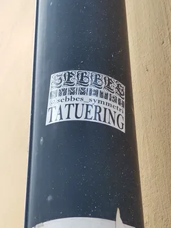 A black and white sticker with the text "Sebbes Symmetri Tatu ering" in a stylized font.  The sticker is attached to a dark grey cylindrical object. The text "Sebbes Symmetri" appears in a smaller font under a larger decorative "Sebbes" title.  The artist's Instagram handle is also included.