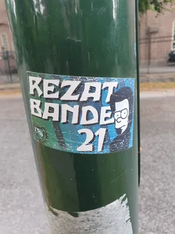 Street sticker A worn sticker featuring the text "REZAT BANDE 21" and an illustration of a person resembling a cartoon character. The sticker is affixed to a green pole.
