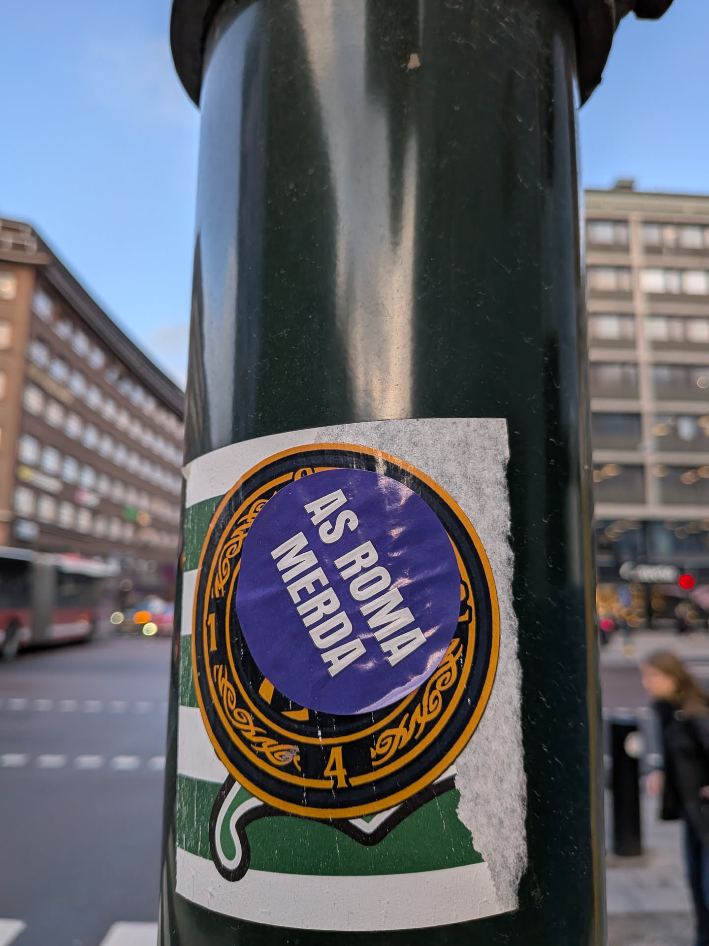 A circular sticker affixed to a dark green pole, showing signs of wear and tear with torn edges. The central design features a vibrant purple circle with white, capitalized text 'AS ROMA MERDA' arranged in an arc. This purple circle is bordered by a thin black ring, followed by a wider gold-colored ring adorned with intricate, swirling patterns reminiscent of Celtic knots. Within this gold ring, the numbers '1927' and '1900' are visible on the left and right sides, respectively. The outermost layer of the sticker, partially visible and torn, consists of vertical green and white stripes.