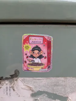 Street sticker This sticker is designed to resemble a box of cereal, with a cute illustration of a girl eating from a bowl of cereal. The sticker has a pink and red color scheme with strawberry accents. It includes text resembling a cereal name and tagline, along with a social media handle.