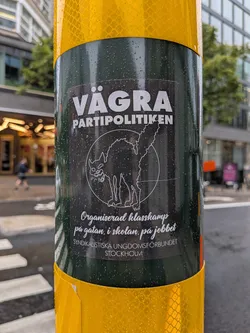 Street sticker A rectangular black sticker with white print, affixed to a wet green pole. The top text reads 'VÄGRA PARTIPOLITIKEN' in bold block letters. Below this is a white line drawing of an arched-back black cat (Sabo-tabby) inside a circle. Under the graphic, cursive text reads 'Organiserad klasskamp på gatan, i skolan, på jobbet'. The bottom line of text reads 'SYNDIKALISTISKA UNGDOMSFÖRBUNDET STOCKHOLM' in a small sans-serif font.
