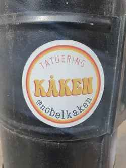 Round sticker with orange, yellow and red stripes.  It says TATUERING (tattoo) in the upper part and KÅKEN in a retro style font in the middle. At the bottom, it shows the Instagram handle @nobelkaken.