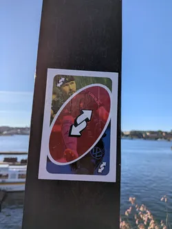 Street sticker The sticker is designed to resemble an UNO Reverse card. The top portion features a close-up of a man's face wearing a black headband and a green and black patterned jersey. The middle section is dominated by a large red oval containing two white arrows forming a circular, reversing symbol. Below this, a blue section shows a portion of a soccer player's leg in pink socks and black shorts with white stripes, and a black Volkswagen logo. The edges of the sticker are white, mimicking the border of an UNO card. There are visible creases and slight peeling at the edges, indicating it is indeed a physical sticker.