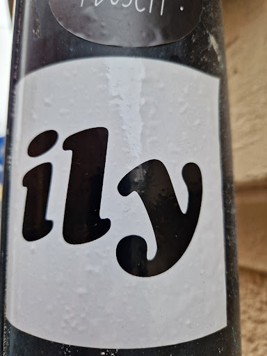 A simple sticker with the abbreviation "ILY" (I Love You) in a stylized font. The sticker is white with black text and is affixed to a dark-colored surface.