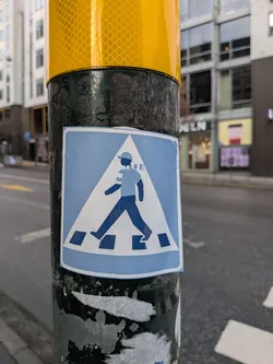 The sticker is a rectangular design with slightly rounded corners, featuring a light blue background and a thin white border. At its center is a large white triangle, reminiscent of a warning sign. Within this triangle, a dark blue, stylized figure of a person is depicted walking from left to right. The figure wears a baseball cap and a short-sleeved shirt with horizontal stripes on the sleeves. Below the figure, several dark blue rectangular blocks represent a crosswalk. The overall color palette is light blue, dark blue, and white. The sticker appears slightly wrinkled and adhered to a textured, dark pole.