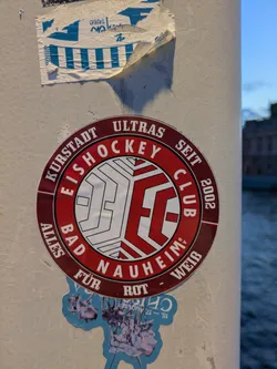 Street sticker The sticker is circular with a dark red border. Inside the border, text reads 'KURSTADT ULTRAS EISHOCKEY CLUB SEIT 2002 ALLES FÜR BAD NAUHEIM ROT - WEISS'. The center features a stylized logo with the letters 'ECC' formed from white lines against a red background, with a geometric, paneled design. Below this main sticker, part of another sticker is visible, featuring a light blue background with metallic-looking figures and text that appears to be a date.