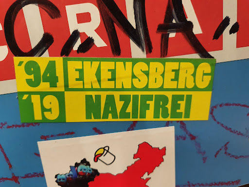 Street sticker A sticker with the text '94 Ekensberg '19 Nazifrei'.  The sticker is yellow and green. The text is in a bold, sans-serif font.