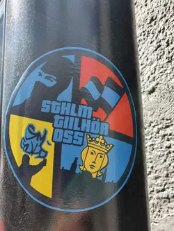 Street sticker Round sticker with a design divided into four sections: a blue section with a stylized flag, a red section with a wavy flag, a yellow section depicting a hand with spray paint, and a blue section with a crowned figure resembling a queen over a cityscape. The text "STHLM TILLHÖR OSS" is prominently displayed.