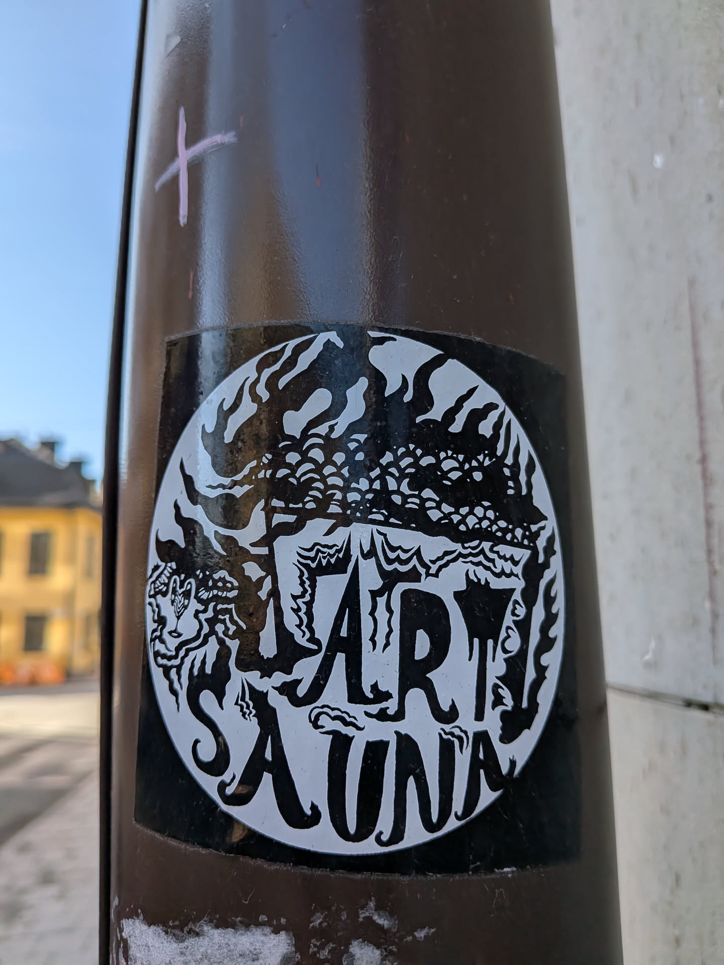 The sticker features a circular black and white design, applied to a larger black square backing, affixed to a brown pole. The circular design showcases white imagery against a black background. The upper half depicts a dense, stylized landscape or cityscape, characterized by numerous small, rounded, organic shapes resembling trees, clouds, or buildings, with flame-like or wavy patterns above them. The lower half prominently displays the text 'ART SAUNA' in a distinctive, somewhat drippy or organic white font, with 'ART' positioned above 'SAUNA'. The overall aesthetic is graphic and illustrative, utilizing strong black and white contrast.
