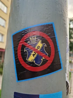 A square sticker affixed to a pole displays a red circle with a diagonal line crossing out an emblem. The emblem is dark blue and gold with what looks like a crown at the top and the letters 'AIS' visible. The sticker has a light blue border.