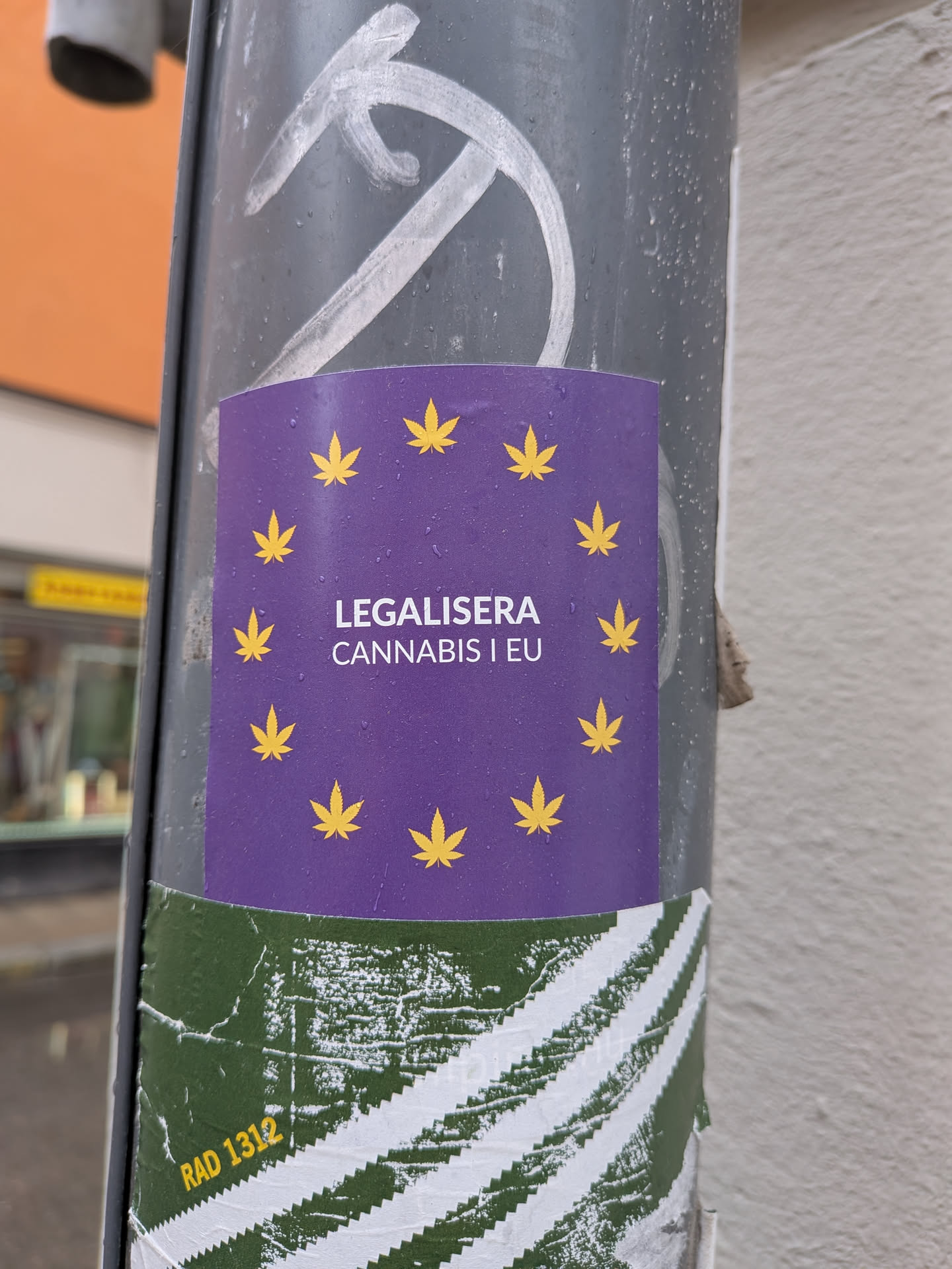 A square-shaped sticker with a deep purple background, featuring twelve yellow cannabis leaves arranged in a circle, mimicking the stars of the European Union flag. In the center, white text reads 'LEGALISERA CANNABIS I EU'. The sticker appears slightly wet with visible water droplets on its surface and is affixed to a grey pole.