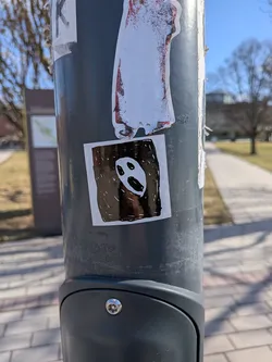 Street sticker The sticker is a roughly square-shaped design with a prominent white border. The central image features a stark black background, against which a minimalist white ghost face is depicted. The face consists of two oval-shaped black eyes and a larger, elongated black mouth, giving it a somewhat surprised or screaming expression. The style appears hand-drawn, with slight imperfections and a glossy finish that reflects light. It is adhered to a grey metal pole.