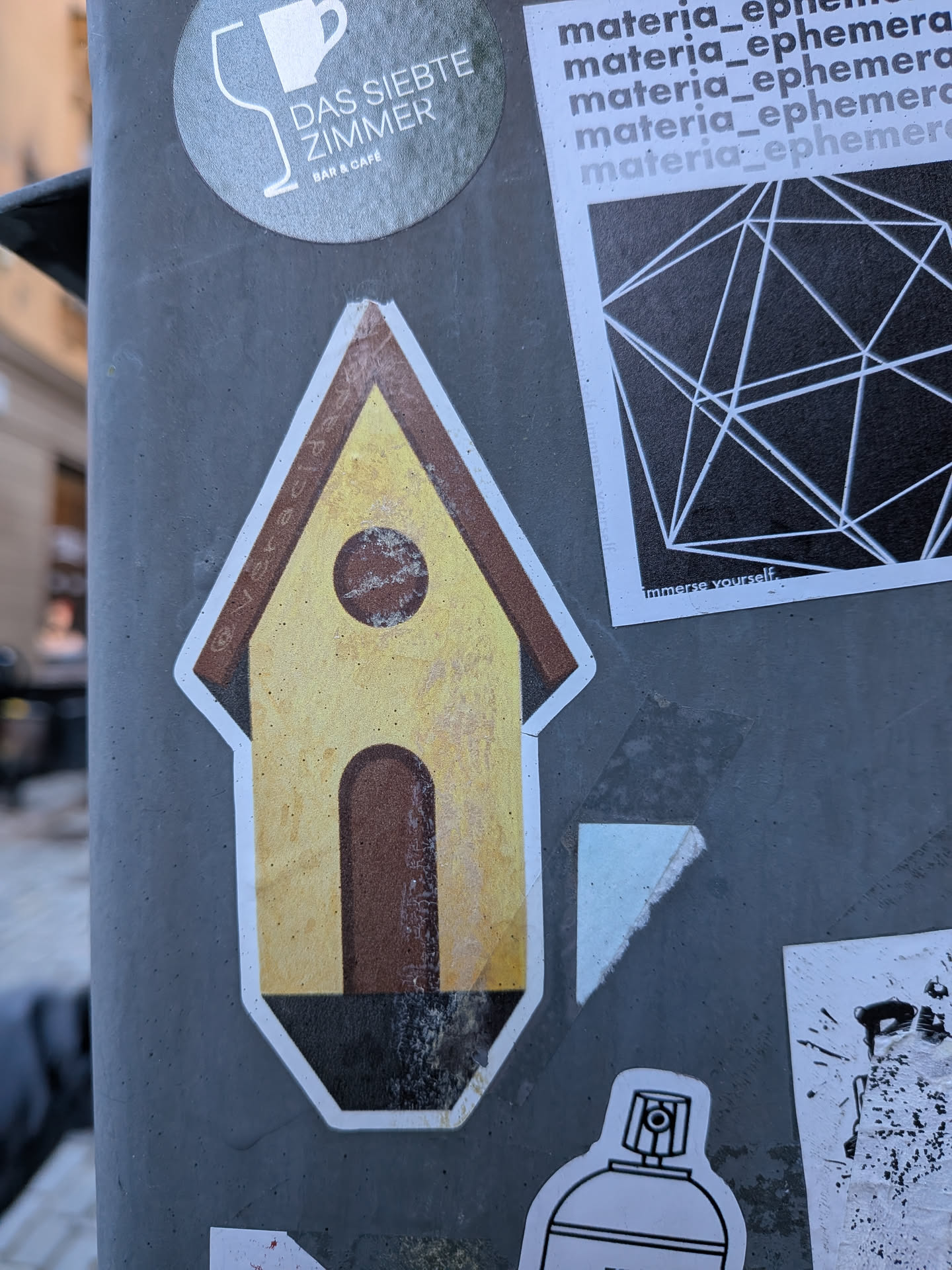 The sticker depicts a stylized birdhouse with a pointed brown roof and a yellow body. It features a small circular opening near the top and an arched entrance below, both rendered in brown. The base of the birdhouse is black. The entire design is outlined in white, and the sticker shows signs of wear and tear, with a slightly textured, faded appearance, particularly on the yellow and brown sections, suggesting it has been exposed to the elements.