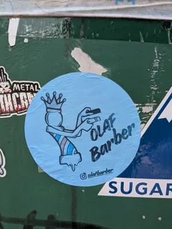 Street sticker A circular light blue sticker featuring a stylized character that combines a barber pole and a crowned figure. The character has arms holding a comb and clippers. The body has diagonal stripes in blue, white, and brown, with a dripping effect at the bottom. The text 'OLAF Barber' is written in a dark blue, hand-drawn style font to the right of the character. An Instagram handle '@olafbarber' is printed at the bottom.