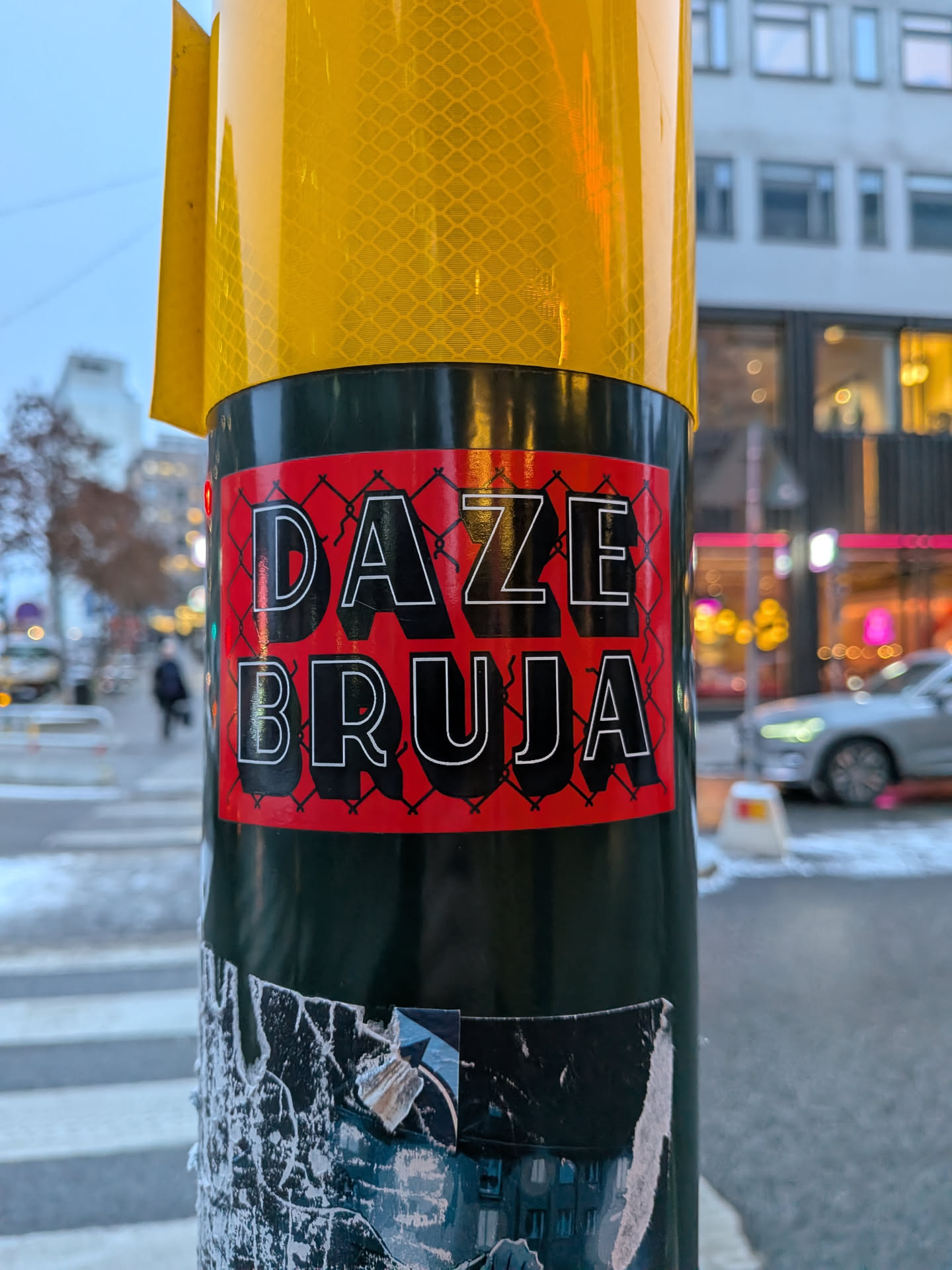 The sticker is rectangular with a vibrant red background. Overlaying the red is a black, diamond-shaped fence pattern. Prominently displayed in large, bold, white-outlined black letters are the words 'DAZE' on the top line and 'BRUJA' on the bottom line. The letters have a distinct 3D effect due to the white outline and black fill. The sticker is affixed to a dark green pole, which is partially obscured by other torn stickers at the bottom.