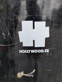 A white sticker with a stylized "HH" logo and the text "HOLLYWOOD.SE" is affixed to a dark, scratched surface. The sticker appears to be made of vinyl and is adhered to the surface unevenly. The background is dark and shows significant signs of wear. The logo is simple and bold, suggesting a brand identity. The overall impression is one of urban street art or branding.