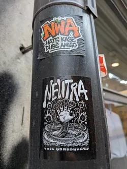 Street sticker A black rectangular sticker featuring the word 'NEUTRA' in a bold, stylized white font at the top. Below the text is a detailed white-on-black illustration of a hand rising from a coiled snake, with each finger depicted as a snake's head. The hand is surrounded by small flaming plants or torches. The bottom of the sticker contains a line of small, highly stylized text.