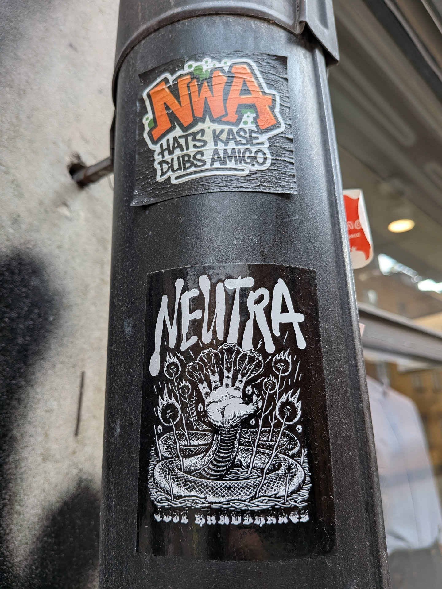 A black rectangular sticker featuring the word 'NEUTRA' in a bold, stylized white font at the top. Below the text is a detailed white-on-black illustration of a hand rising from a coiled snake, with each finger depicted as a snake's head. The hand is surrounded by small flaming plants or torches. The bottom of the sticker contains a line of small, highly stylized text.