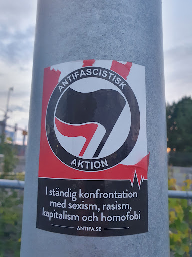 Street sticker A sticker with the Antifa symbol and the text "Antifascistisk Aktion" in Swedish. The text below translates to: "In constant confrontation with sexism, racism, capitalism, and homophobia."  A website address, antifa.se, is also included.