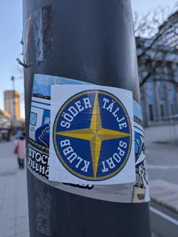 Street sticker A circular sticker featuring a prominent four-pointed yellow star or compass rose design at its center, set against a deep blue background. The star has a small yellow square in its middle. Encircling the central design is a blue band with white capital letters that read 'SÖDER TÄLJE' at the top and 'SPORT KLUBB' at the bottom, following the curve of the circle. A thin yellow border outlines the blue band. The sticker has a slightly worn appearance, indicating it's a physical adhesive.