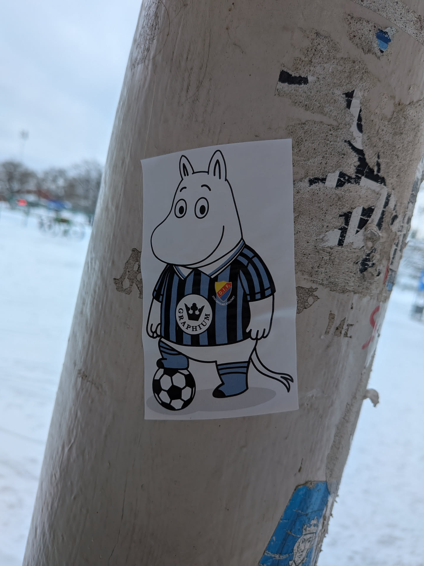 Street sticker The sticker features a white, cartoon Moomintroll character, outlined in black, standing with a classic black and white football at its left foot. It is wearing a black and light blue striped football jersey with light blue shorts and socks. On the right side of its chest (viewer's left), there's a circular white logo with a black crown and the word "GRAPHIUM" in black text below it. On the left side of its chest (viewer's right), there's another circular logo with "D.I.F." at the top, "STOCKHOLMS STOLTHET" at the bottom, and a yellow and red shield emblem in the center. The Moomintroll has a friendly expression with large black eyes. The sticker is rectangular with a white background.