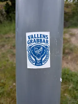 Street sticker A rectangular white sticker with blue print. It features a hooded figure holding flares, framed by a laurel wreath and a shield containing the text 'IFK LIDINGÖ'. The words 'VALLENS GRABBAR' are arched at the top, and '2025' is printed at the bottom.