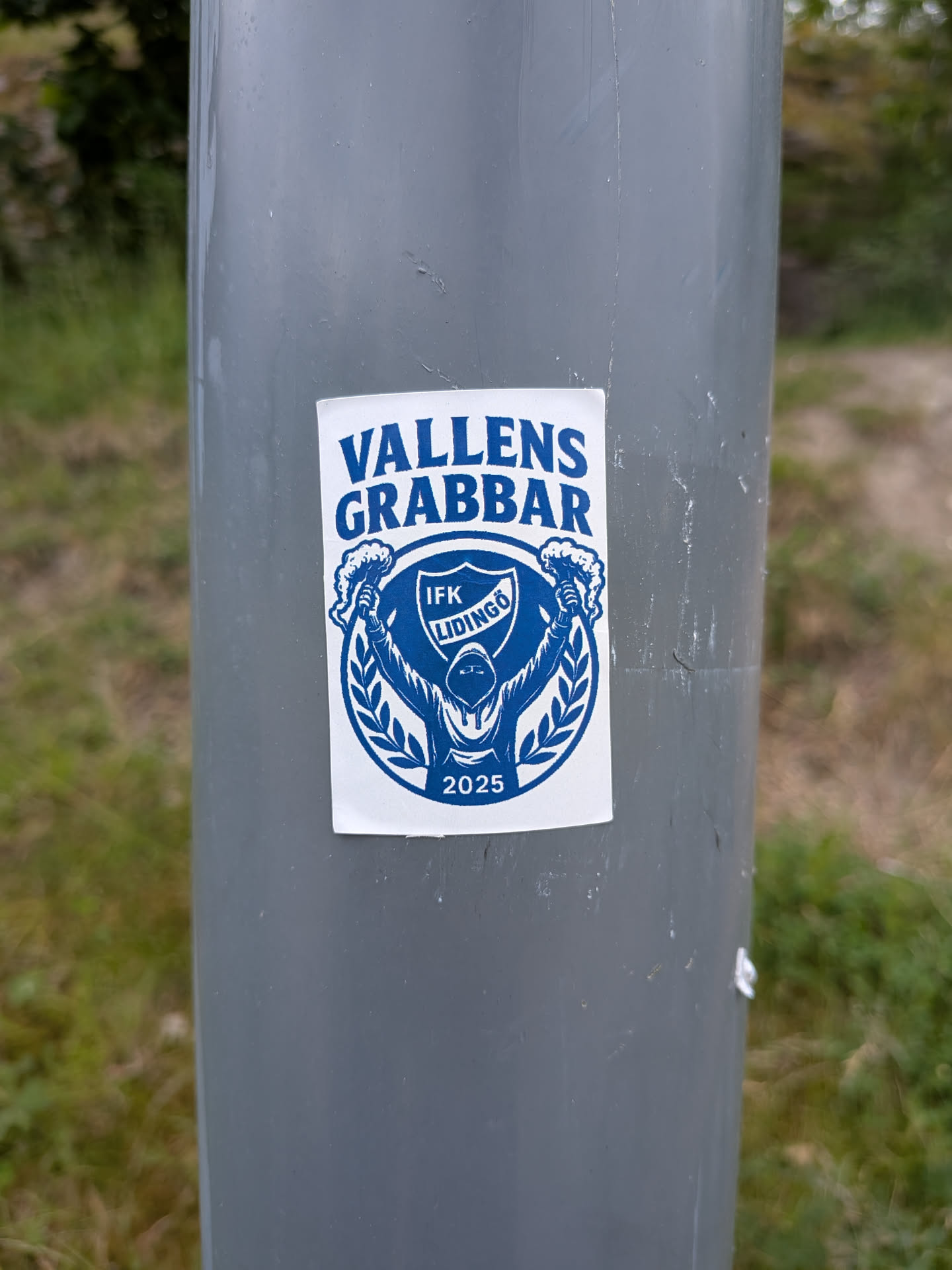 A rectangular white sticker with blue print. It features a hooded figure holding flares, framed by a laurel wreath and a shield containing the text 'IFK LIDINGÖ'. The words 'VALLENS GRABBAR' are arched at the top, and '2025' is printed at the bottom.