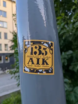Street sticker Rectangular sticker with a yellow center and black border decorated with bubbles. Features the number '135', 'Biblioteksgatan 8, Stockholm', 'AIK', and 'Styrka · Stolthet · Tradition' in black.
