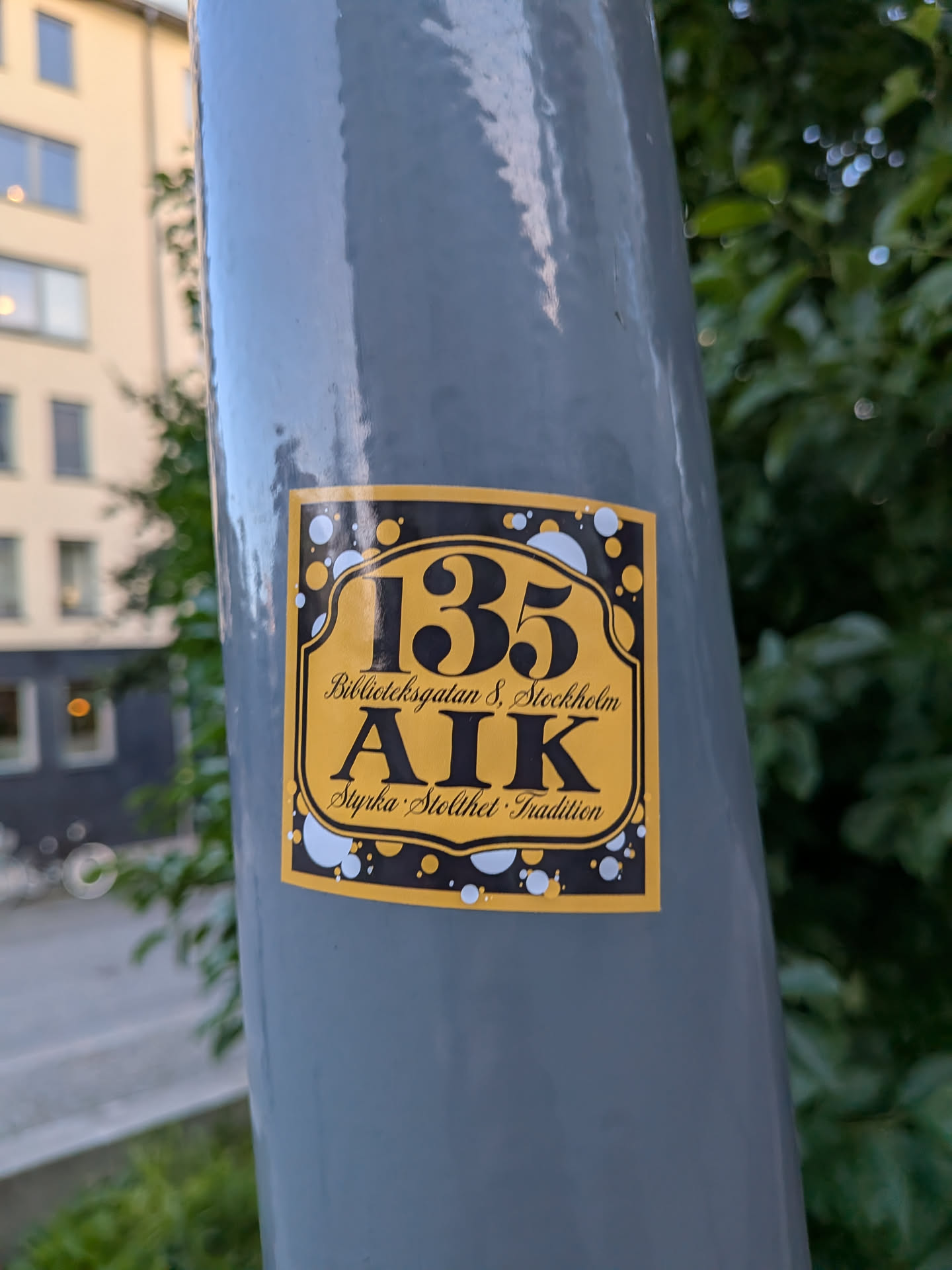Rectangular sticker with a yellow center and black border decorated with bubbles. Features the number '135', 'Biblioteksgatan 8, Stockholm', 'AIK', and 'Styrka · Stolthet · Tradition' in black.