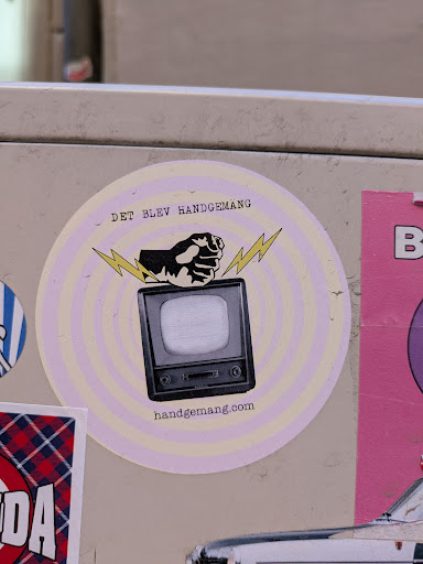 The sticker is a circular design featuring a vintage television set with a fist punching down on it, accompanied by lightning bolts. The background consists of concentric circles in a pale lavender and white color scheme. The text "DET BLEV HANDGEMANG" is arched above the image, and "handgemang.com" is printed below.
