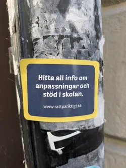 A blue sticker with yellow borders is affixed to a pole, displaying the text 'Hitta all info om anpassningar och stöd i skolan' and the website 'www.rattpariktigt.se'. It is advertising information about school adaptations and support.