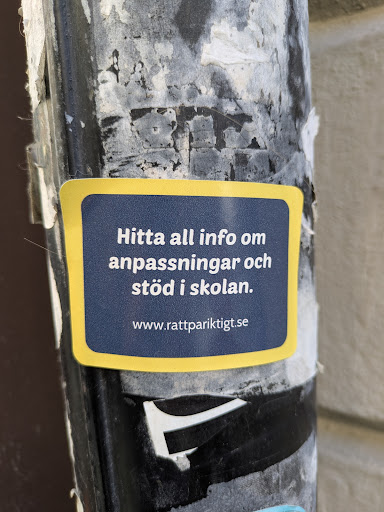 A blue sticker with yellow borders is affixed to a pole, displaying the text 'Hitta all info om anpassningar och stöd i skolan' and the website 'www.rattpariktigt.se'. It is advertising information about school adaptations and support.