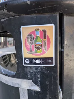 Street sticker The sticker features a stylized illustration set against a warm, light orange background with a prominent pink circular element behind the main objects. From left to right, it depicts a vintage-style silver microphone on a stand, a green bottle with a light blue neck and a label that reads 'KRIMINAL TANGO', and a brown-handled knife with a silver blade. Below this illustration, there's a black bar with a white Spotify logo on the left and a Spotify 'scan code' (a series of vertical bars) on the right. The sticker has a thin white border around its rectangular shape with slightly rounded corners.