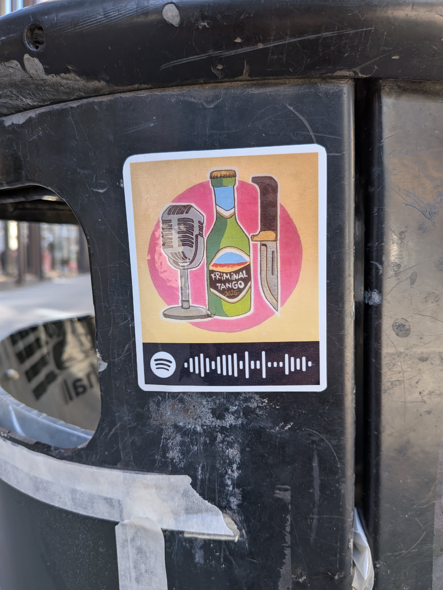 The sticker features a stylized illustration set against a warm, light orange background with a prominent pink circular element behind the main objects. From left to right, it depicts a vintage-style silver microphone on a stand, a green bottle with a light blue neck and a label that reads 'KRIMINAL TANGO', and a brown-handled knife with a silver blade. Below this illustration, there's a black bar with a white Spotify logo on the left and a Spotify 'scan code' (a series of vertical bars) on the right. The sticker has a thin white border around its rectangular shape with slightly rounded corners.