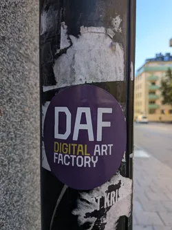 Street sticker The sticker is circular and predominantly purple. In the center, it features the large, bold white letters 'DAF'. Below this, in smaller yellow letters, it reads 'DIGITAL ART', and further below, in smaller white letters, 'FACTORY'. The sticker appears to be placed on a dark, weathered pole, with other torn and faded stickers partially visible around it.