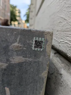 Street sticker The sticker is square-shaped with a white background and a green border. It features a black QR code in the center, surrounded by a repeating pattern of small, stylized green icons that resemble buildings or pagodas. The sticker appears to be adhered to a rough, textured surface, possibly concrete or stone, with some dirt and wear visible.
