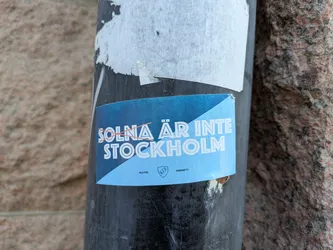 The sticker is primarily light blue with white text and a shield emblem. The text "SOLNA ÄR INTE STOCKHOLM" is prominently displayed across the top portion, with an orange line crossed through "SOLNA". Below this, the word "STOCKHOLM" is written in larger letters. At the bottom, there are smaller text elements "ALLTID." and "GAVSETT." flanking a shield with "D.I.F." inside. The sticker has a slightly curved, possibly oval or rounded rectangular shape, and is affixed to a dark, cylindrical surface. The edges of the sticker show some wear and tear, indicating it has been applied for some time.