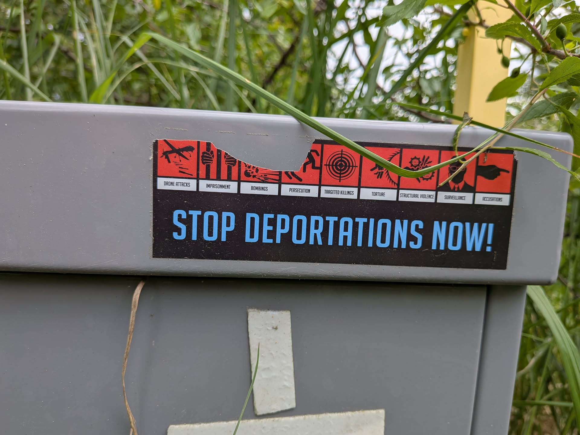 A rectangular sticker with a black bottom half featuring the blue text 'STOP DEPORTATIONS NOW!'. The top half displays a row of red icon squares depicting state violence and human rights abuses, each with a white label underneath. The sticker is partially torn at the top.