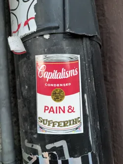 Street sticker The sticker is designed to mimic the iconic Campbell's Soup can label, but with a critical message about capitalism. The top section features the word "Capitalism's" in a stylized, cursive font, followed by "CONDENSED" in a block font. Below this, a gold circular emblem with an embossed image is present. The main body of the sticker displays "PAIN & SUFFERING" in large, bold, red capital letters. The bottom of the sticker has a wavy banner with the word "SUFFERING" in a decorative, gold-colored font. There are also small, colorful heart icons and some numbers printed in the lower right corner of the sticker.