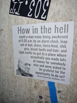 Street sticker A rectangular white sticker featuring a black-and-white portrait of author Charles Bukowski alongside a printed quote from his 1975 novel 'Factotum' questioning the routine of modern employment.
