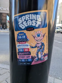 A rectangular sticker with slightly rounded corners, adhered to a black pole. The background is a warm peach/orange hue. At the top, the event name 'SPRING BEAST' is prominently displayed in large, bubbly white letters against a wavy blue background, with '2026' in pink below it. The central figure is a cartoonish, pink-skinned creature with small horns, pointed ears, a wide grin showing many teeth, and a single large blue eye on its forehead. This character is clad in a blue and pink robotic suit, pointing to the left. To the left of the character, various event details are presented in distinct text blocks: 'MAY 14-17 FESTIVAL' in blue and white, 'LIVE MUSIC' in red, 'LOOPTROOP ROCKERS' in blue and red, and 'LINDA PIRA + MANY MORE!' in pink and white. Further down, 'GRAFFITI' and 'STREET ART' are highlighted in dark blue and pink. The bottom section lists 'ART MARKET', 'FOOD TRUCKS', 'DJ SETS', and 'WORK SHOPS' in smaller text. A dark purple/blue cityscape silhouette forms the base of the design. The bottom right corner features a 'TICKETS' label above a QR code. The overall aesthetic is energetic, colorful, and playful.