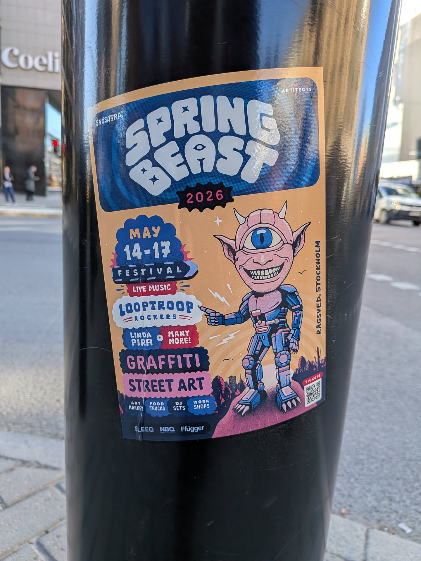 A rectangular sticker with slightly rounded corners, adhered to a black pole. The background is a warm peach/orange hue. At the top, the event name 'SPRING BEAST' is prominently displayed in large, bubbly white letters against a wavy blue background, with '2026' in pink below it. The central figure is a cartoonish, pink-skinned creature with small horns, pointed ears, a wide grin showing many teeth, and a single large blue eye on its forehead. This character is clad in a blue and pink robotic suit, pointing to the left. To the left of the character, various event details are presented in distinct text blocks: 'MAY 14-17 FESTIVAL' in blue and white, 'LIVE MUSIC' in red, 'LOOPTROOP ROCKERS' in blue and red, and 'LINDA PIRA + MANY MORE!' in pink and white. Further down, 'GRAFFITI' and 'STREET ART' are highlighted in dark blue and pink. The bottom section lists 'ART MARKET', 'FOOD TRUCKS', 'DJ SETS', and 'WORK SHOPS' in smaller text. A dark purple/blue cityscape silhouette forms the base of the design. The bottom right corner features a 'TICKETS' label above a QR code. The overall aesthetic is energetic, colorful, and playful.