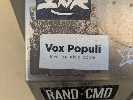 A rectangular white sticker with the text 'Vox Populi Investigando al poder' in black font. The sticker is affixed to a metallic surface, likely a public utility box or similar structure. The surrounding area shows signs of wear and other stickers.
