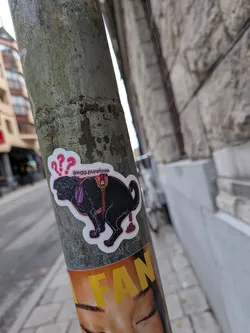 The sticker features a black dog, possibly a Labrador or similar breed, in the process of defecating. The dog is wearing a purple collar and a red harness with gold buckles. Above the dog's head are three pink question marks, suggesting confusion or surprise. The sticker has a white border and is placed on a textured, grey pole. The overall style is illustrative and somewhat cartoonish.